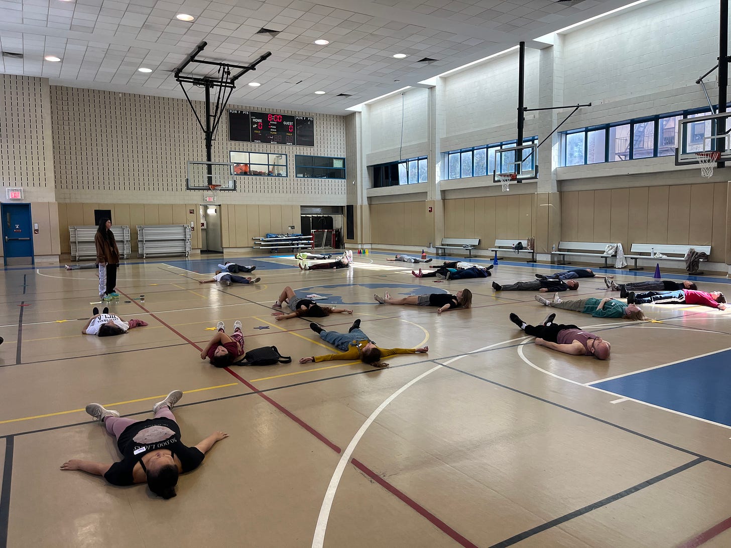 In a light-filled gymnasium, about 15 people lie on their backs on the floor, scattered throughout the space.