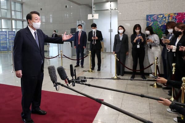 President Yoon Suk Yeol, wearing a white face mask and a dark suit and standing on a red carpet, addresses journalists. Some are extending microphones in his direction.