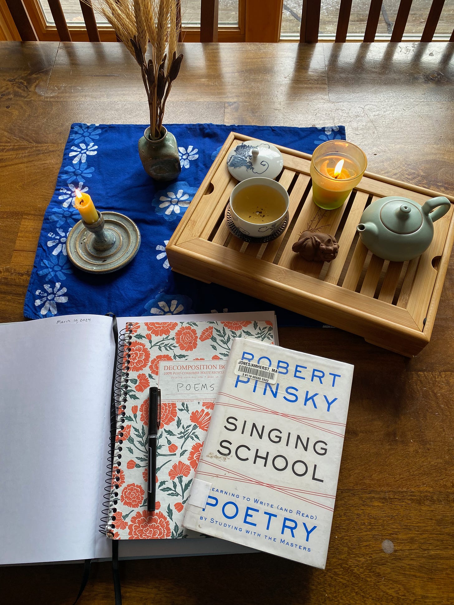 My poetry school setup: a stack of notebooks, Robert Pinsky’s Singing School, a blue cloth placemat with a candle and a vase of dried flowers, and a tea tray with teapot, cup, and tea animal.
