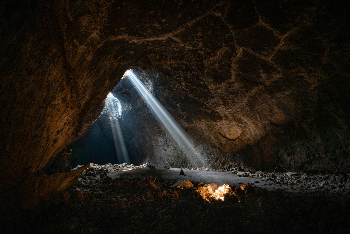 Photo from inside a cave. Walls are brown and floor is dark. There are 2 holes in the middle of the photo where light shines in as 2 independent beams. One shines on the ground and illuminates the rocks in a bright yellow glow.
