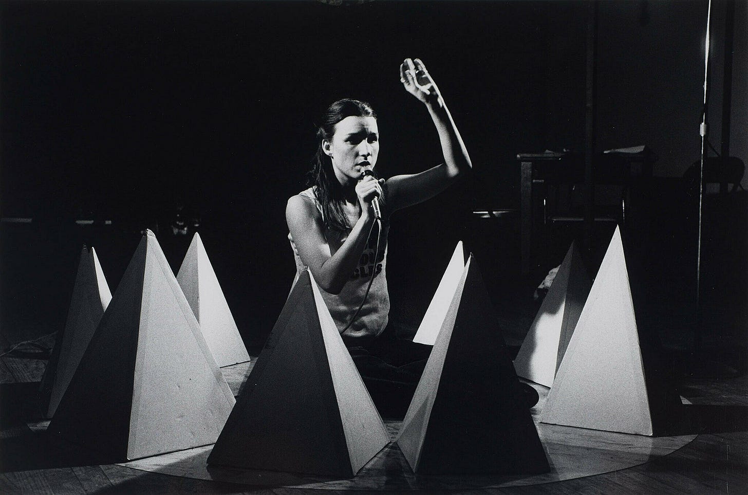 A woman kneels on a stage surrounded by sculptural objects.