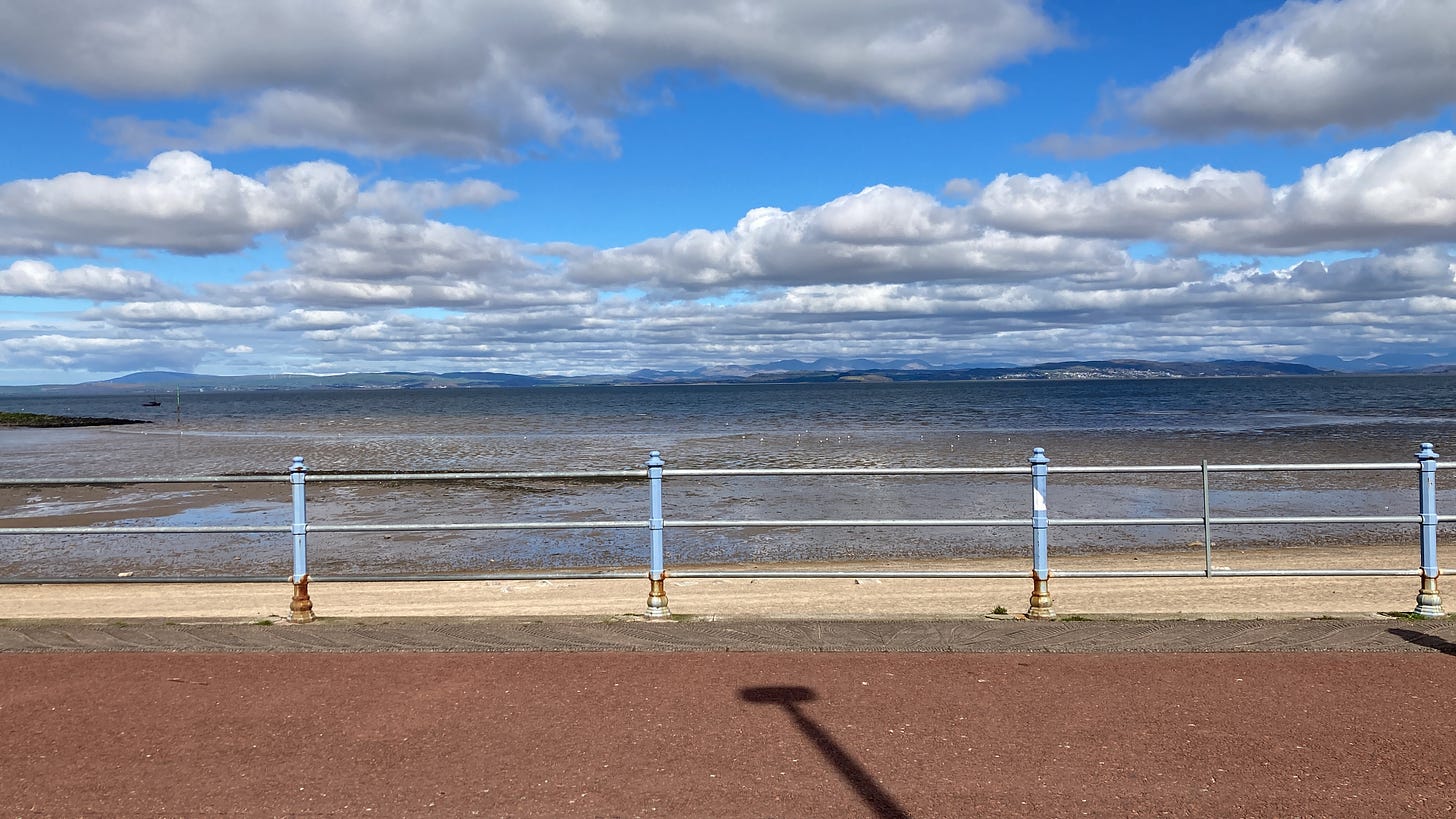 A cloudy sky with visible peaks across the bay on the horizon. The tide is out, framed by the railings of the Promenade, peeling blue paint with patches of rust. There is a dark shadow from a lamp post in the foreground.