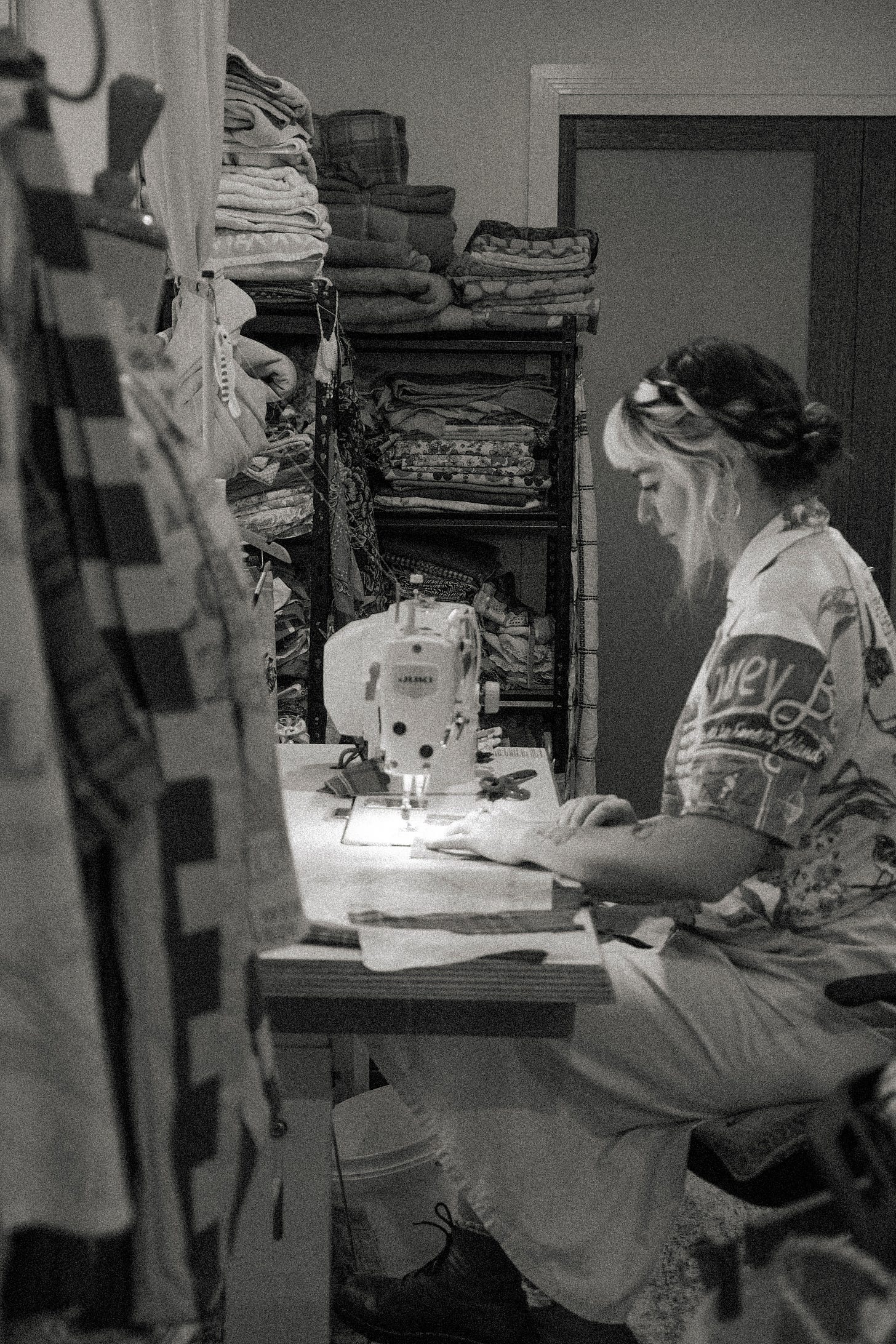 a photo of Clare from House of Clare sewing on her sewing machine. She is sitting at a table with her fabrics sitting next to her on a big shelf. She is wearing one of her beautiful button up shirts.