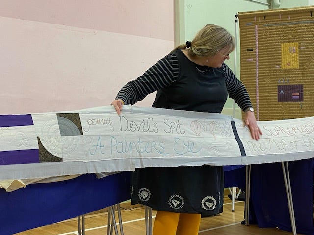 A woman holds a long strip of quilting pointing out parts of it.