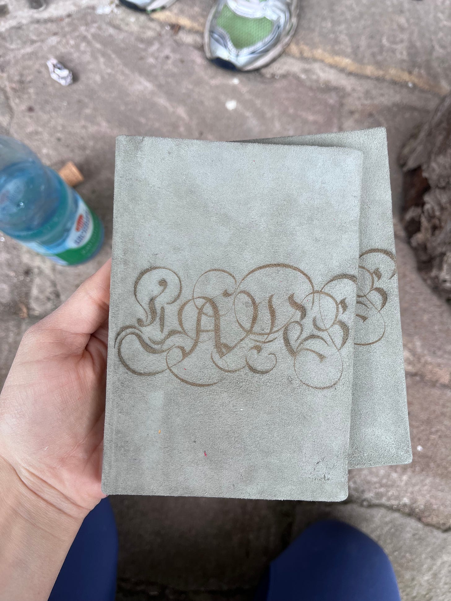 A POV shot of two A5 books, grey suede, one on top of another face up. The title is laser cut in large cursive captial letters, spelling LAMB. A hand holds the books and in the background you can see a water bottle and some shoes.