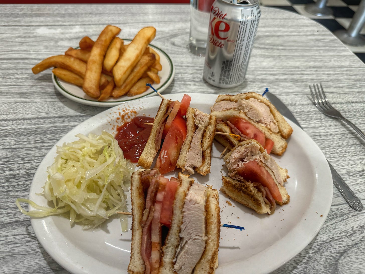 Tabletop photo of a dinner plate with a club sandwich, cole slaw, and ketchup on it. In the background there is a dish of french fries and a can of Diet Coke. The ketchup on the plate is spattered in a way that suggests the ketchup bottle was running out.