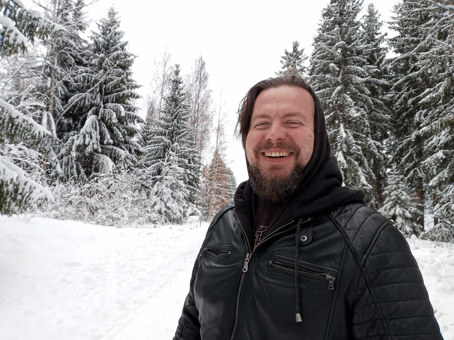 A photograph of Jakub, smiling widely as he's standing surrounded by snow-covered trees and piles of snow on the road.
