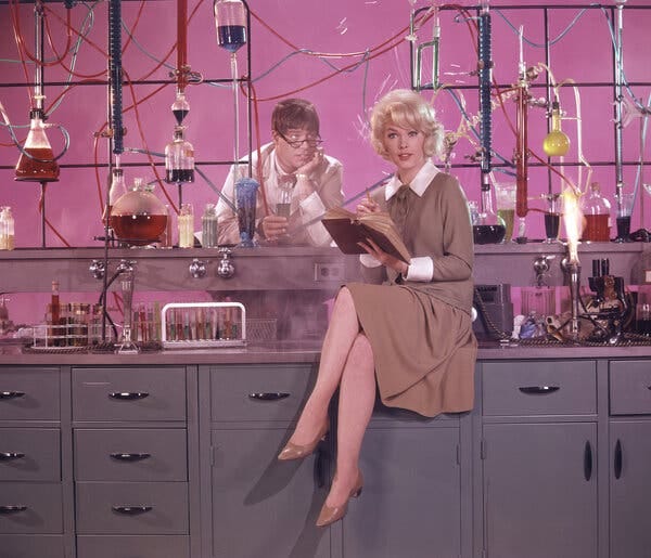 A color photo of Ms. Stevens -- blond, in a brown dress and sitting on a laboratory counter with her legs crossed. Behind her is Jerry Lewis, with a comical expression, leaning over the counter with his eyeglasses perched on the end of his nose