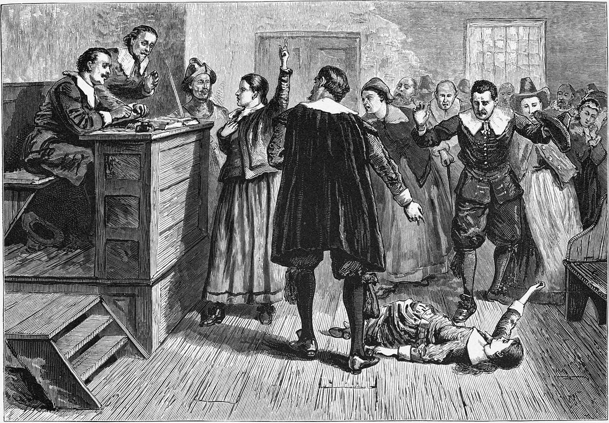 An engraved illustration of the Salem witch trials. One woman stands with her hand raised, as though declaring her experience, and another lays stricken on the floor. Two keepers of justice look on with perturbed concern, and the onlookers are wary.