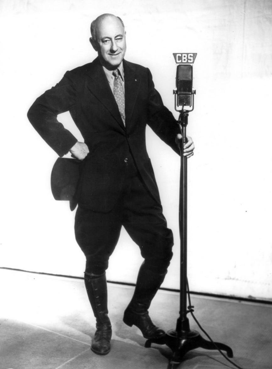 Cecil B. DeMille posed at a microphone, smiling slightly, wearing a blacl riding outfit, holding a black hat at his waist