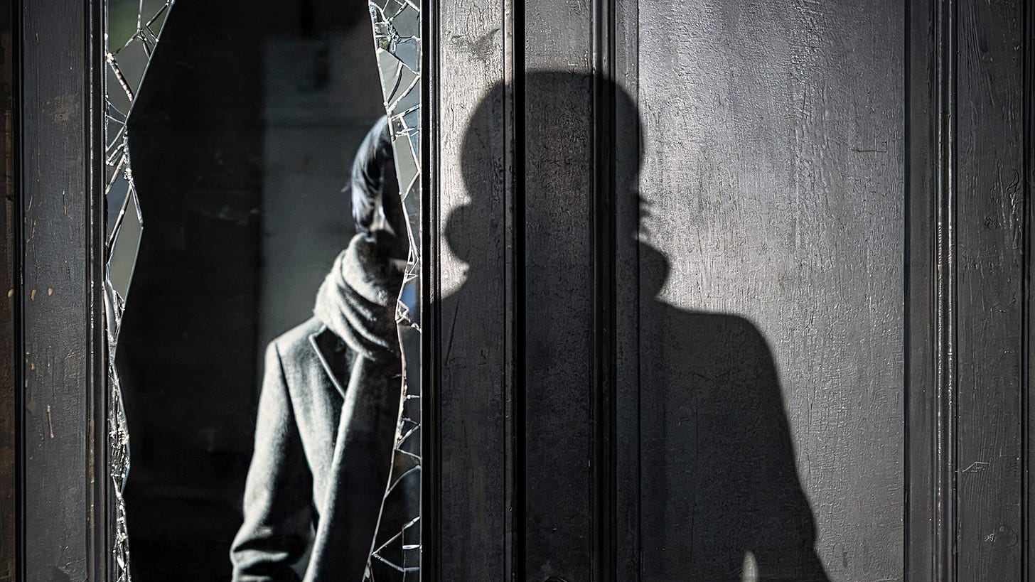 A haunting black and white image shows a broken glass panel in a dark door, with a shadowy figure visible through the jagged break. The person's shadow looms larger on the adjacent door panel, creating a dual presence that echoes the poem's themes of watching and being watched, fragmented identity, and the persistence of trauma's witness. The broken glass mirrors the poem's climactic moment of mirror-smashing, while the shadow suggests both vulnerability and emerging strength.