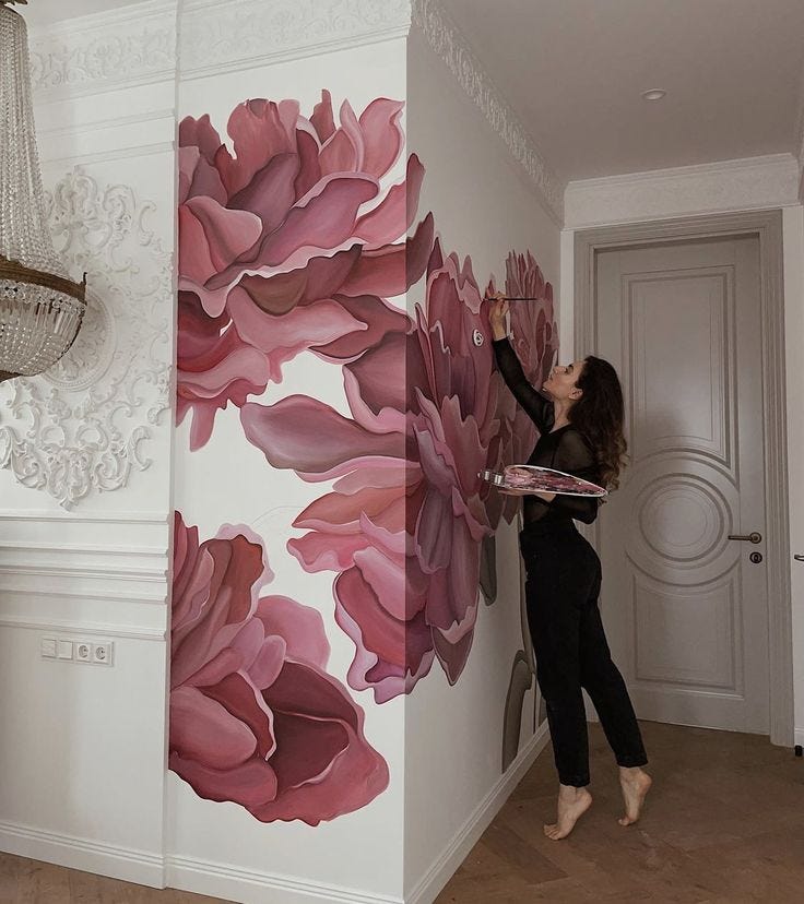 This may contain: a woman painting a wall with pink flowers on it in a white room next to a chandelier