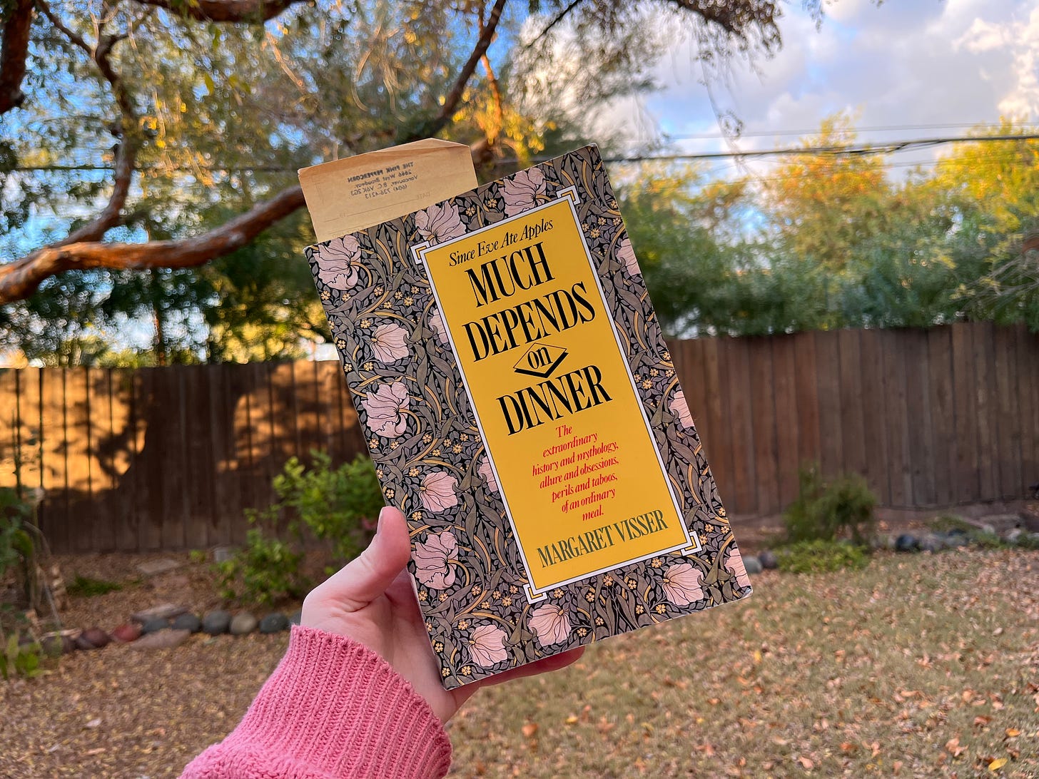 my left hand holding a copy of Much Depends on Dinner: The extraordinary history and mythology, allure and obsessions, perils and taboos, of an ordinary meal (1986) by Margaret Visser in my backyard