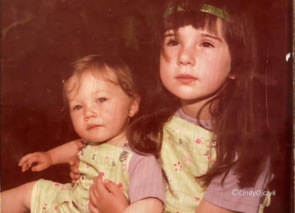 A toddler sits on the lap of a slightly older girl both dressed in violet shirts and green coveralls