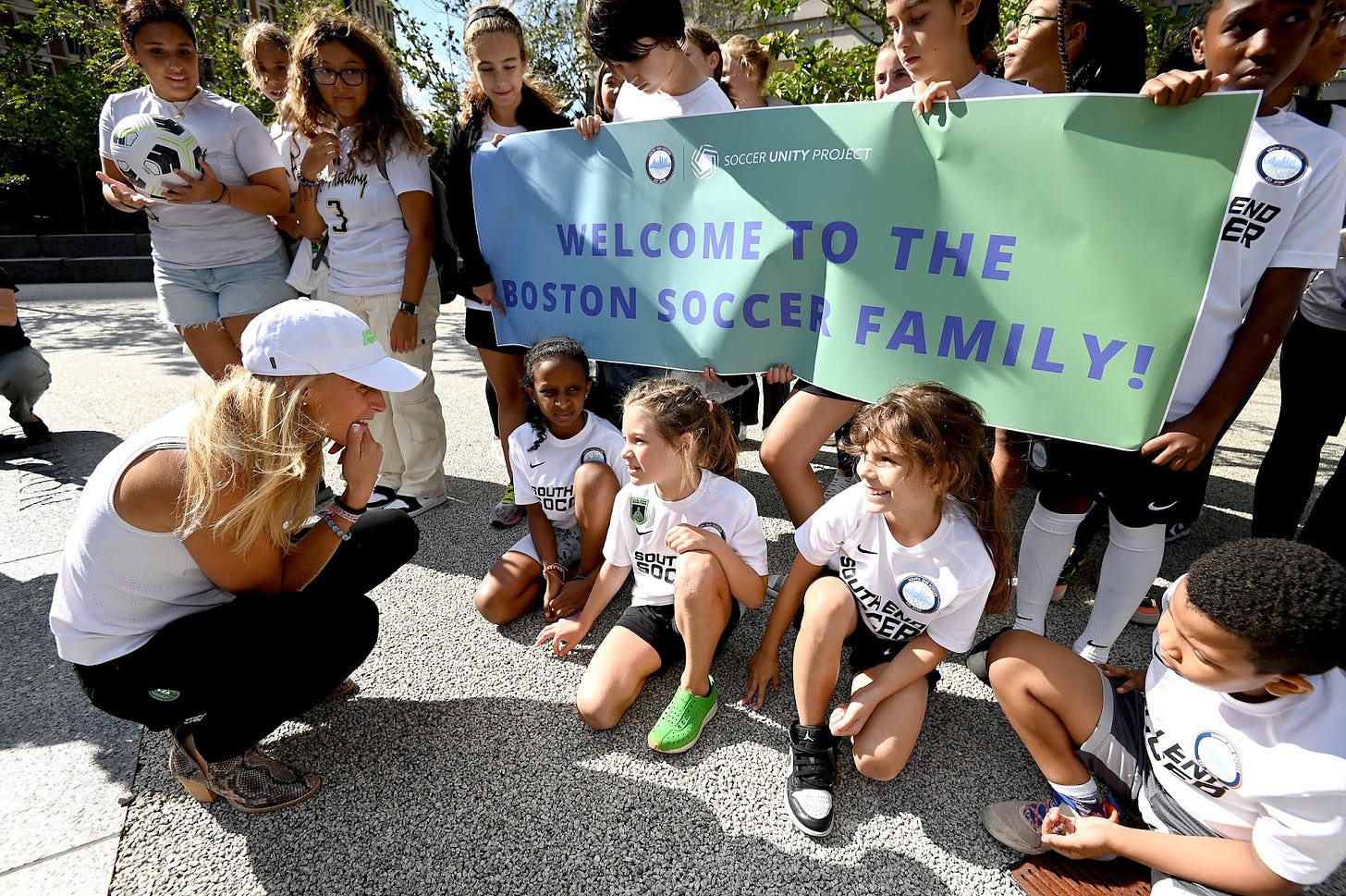Christine Lilly kneels down to speak with young children wearing South End Soccer shirts while others hold a banner that reads "Welcome to the Boston soccer family!"