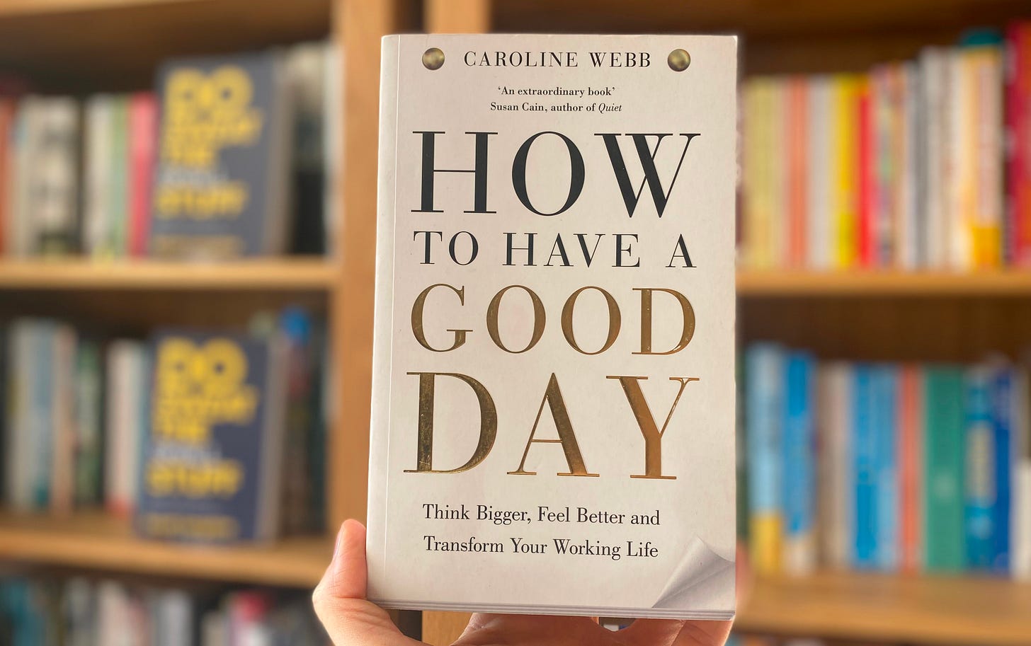 A photo of the book, How to Have a Good Day by Caroline Webb, in front of a blurry set of bookcases. The book has a white cover in the style of a tear off pad, with the bottom right hand corner appearing slightly lifted. The title is in black and gold capital letters, and the subheading: Think Bigger, Feel Better and Transform Your Working Life is at the bottom.