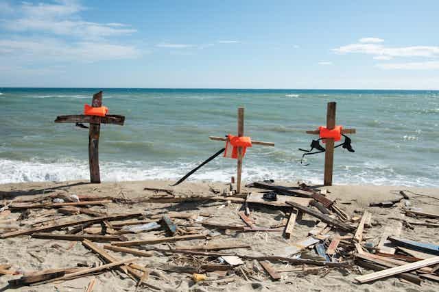 Crosses made of pieces of wood, and other pieces lying on a beach at the water's edge.