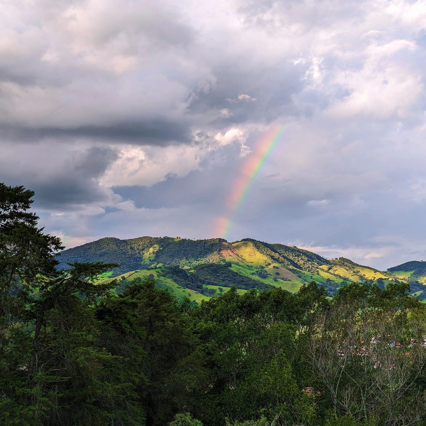 A rainbow appears in the distance, rising from the Mantiqueia mountain range in southern Minas Gerais, into a sky with white and grey skies.