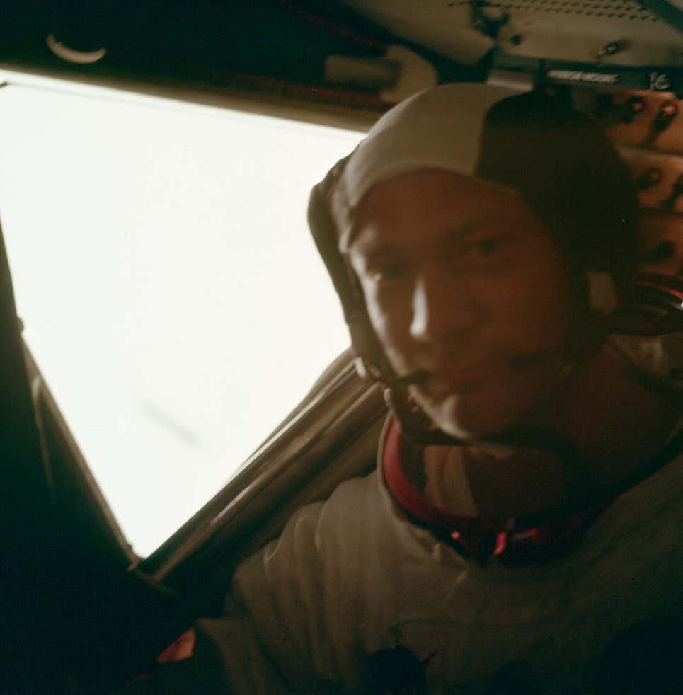 A blurry photo of buzz aldrin in space suit inside the lunar module, a bright triangular window glowing behind him.