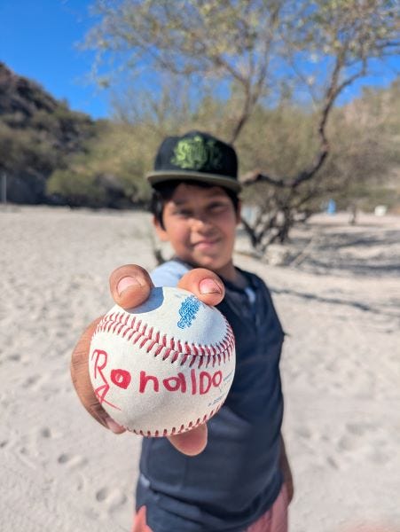 a young boy on a sandy beach, holding a baseball out in front of him with a pitcher's grip. The ball says Ronaldo in childish red printing.