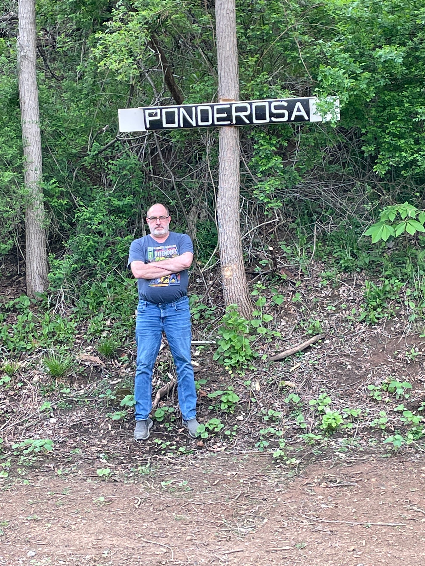 Brian Keene standing in a wooded area in front of a sign. 