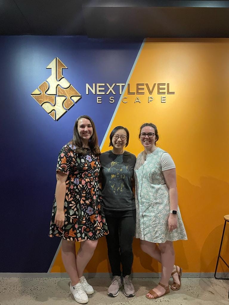 Three women standing in front of a wall that's half blue and half yellow, below a logo of a puzzle and the words Next Level Escape Room