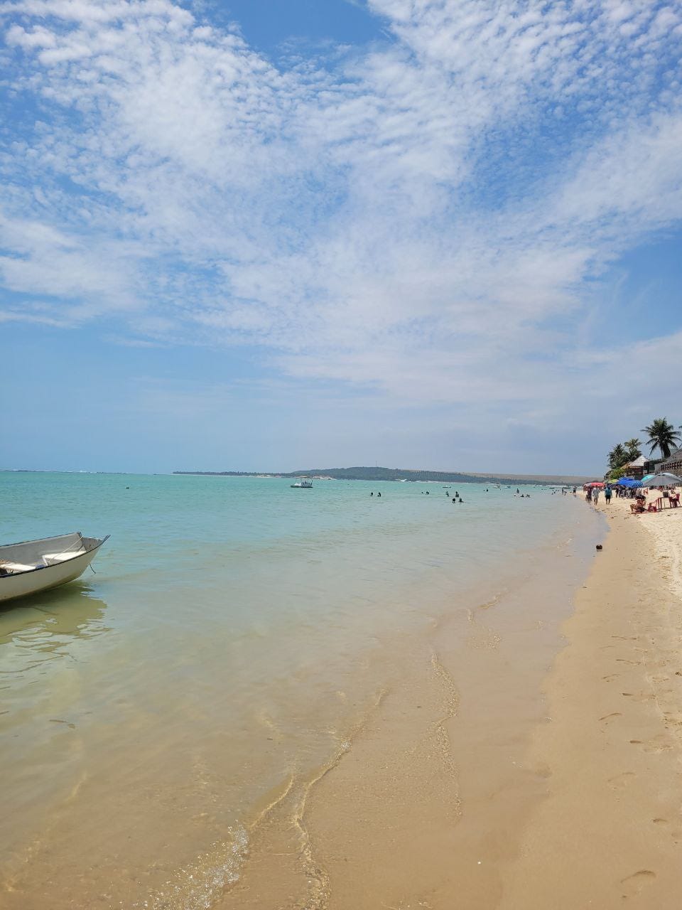 Uma vista de praia com mar muito azul, alguns guarda-sóis e barcos e bem ao fundo uma área verde.