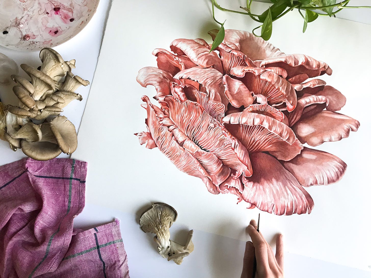 A picture from above of the finished pink oyster mushroom, with real mushrooms laying to the side of the painting, and the paint palette in the top right corner. The painters hand is reaching into the frame with a paintbrush. 