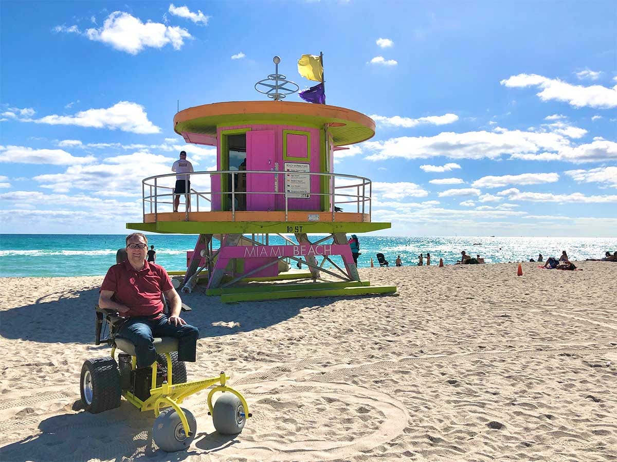 John seated on a beach wheelchair in front of a life guard stand.
