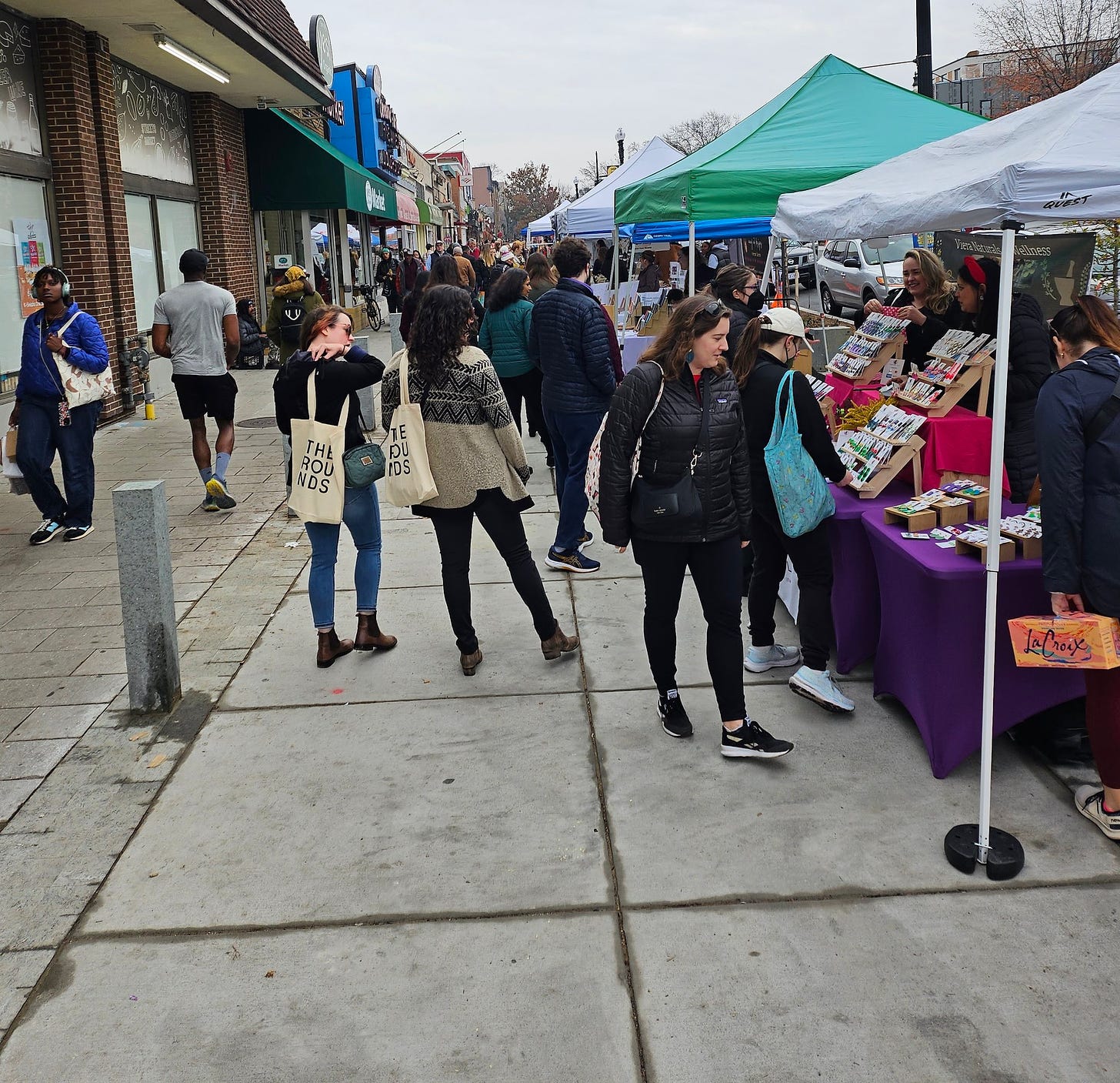 Photo of the Cleveland Park Promenade standing in front of streets market looking south. You can see the streets market awning on the top left. There's a large crowd of people visible all the way up the horizon, at least 50 clearly visible. On the left, are a line of tents with small businesses. The whole area is crowded. They are standing on the new Streetscape design. 