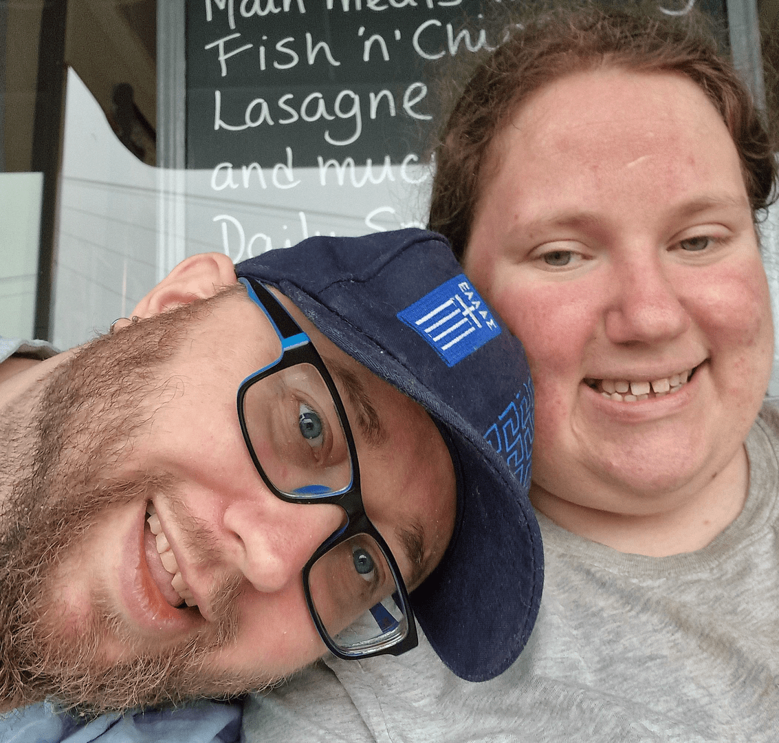 A photo of me (left) and Cuddles (right). I’m wearing a blue cap with the Greek flag on, glasses, and a beard, and smiling at the camera. Cuddles wears her brown hair pulled back, and a grey shirt, and is also smiling at the camera.