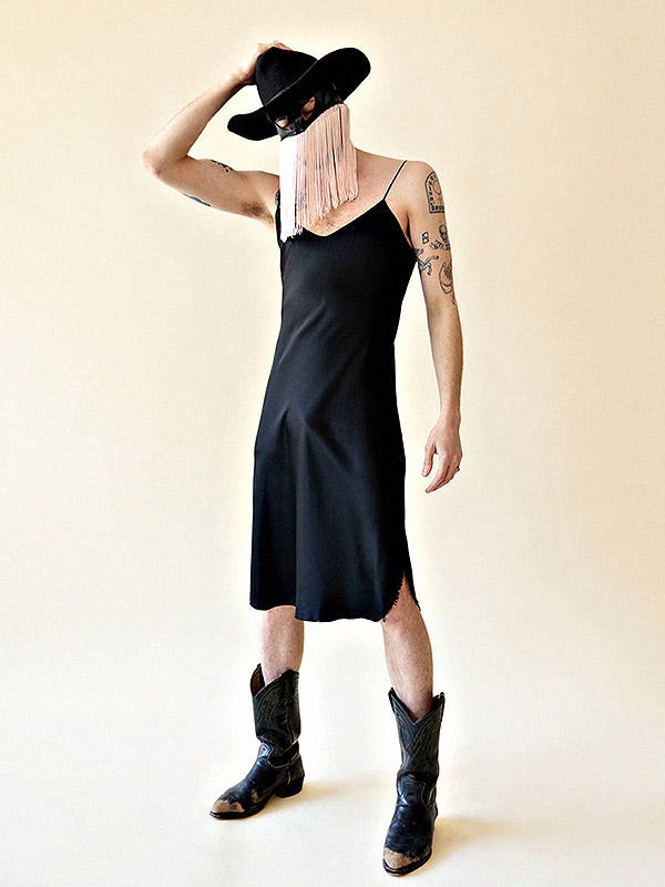 Orville peck in a black spaghetti strap dress, black mask with pink fringe, black cowboy hat, and black boots. His hand is on his hat.