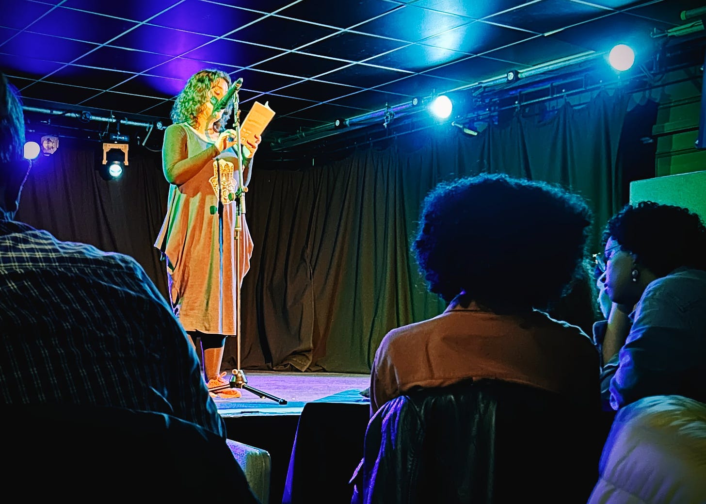 An image capturing a live performance by Sara Shaarawi, who is presenting an excerpt from her play 'Sister Radio' at The Lemon Tree venue. The performer, positioned on stage with a microphone stand, is reading from a script. The stage lighting casts a vibrant glow around her, accentuating her presence. In the foreground, the audience is attentively listening, with some members visible from the back, creating an intimate atmosphere of engagement between the performer and her audience.