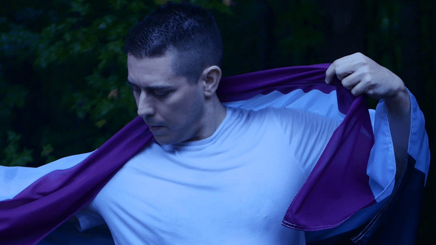 The author drapes the asexual flag (black, gray, white, and purple stripes) over his shoulders. He looks down to the side and is standing in front of a forest in the rain.