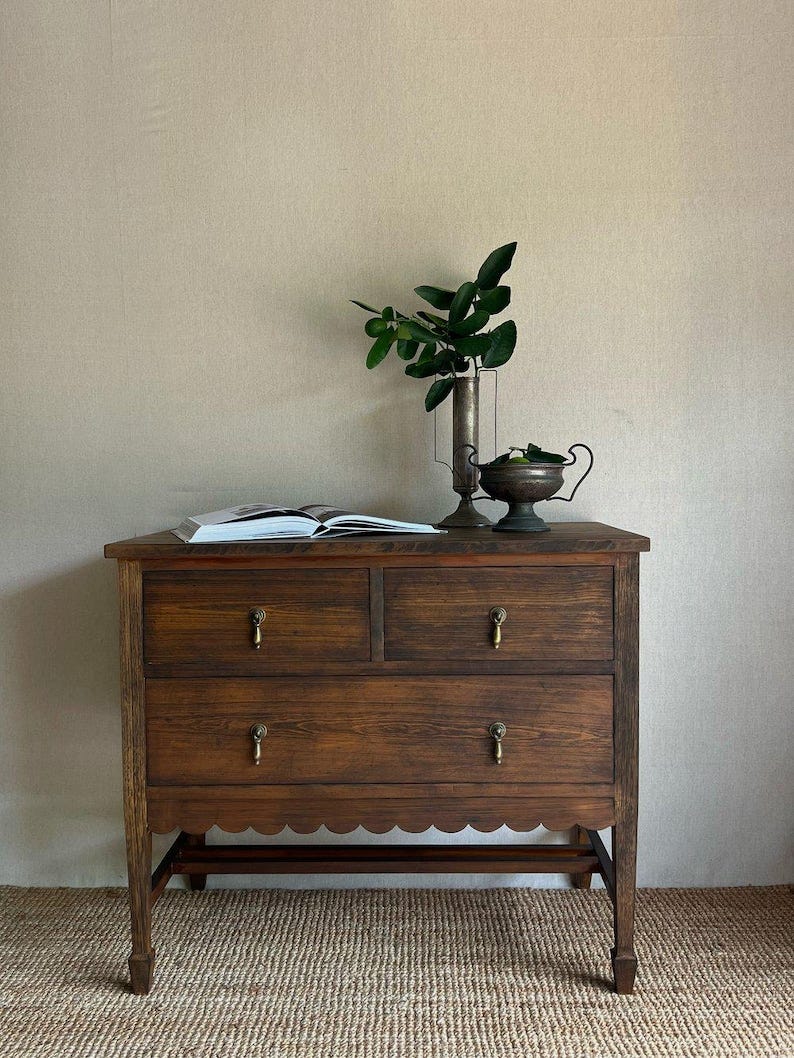 May include: A three-drawer wooden dresser with a scalloped bottom edge. The dresser is dark brown and has brass knobs. The dresser is sitting on a brown woven rug in front of a beige wall.