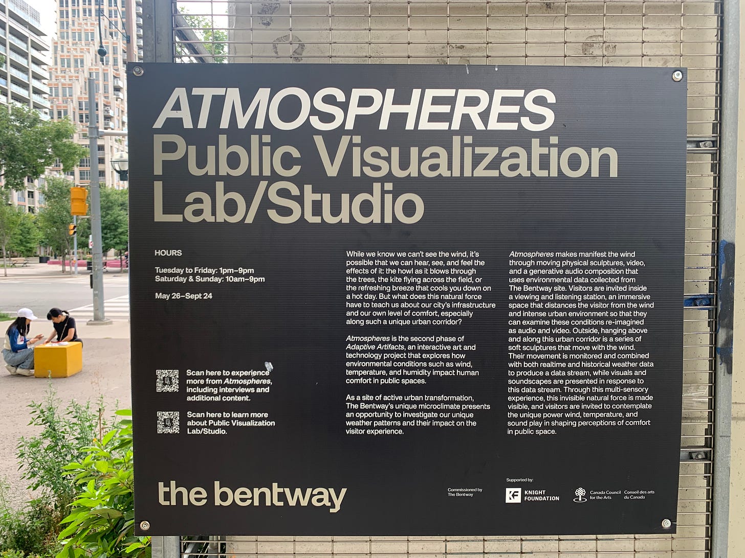 Sign that says: "ATMOSPHERES, Public Visualization Lab/Studio. While we know we can't see the wind, it's possible that we can hear, see and feel the effects of it: the howl as it blows through the trees, the kite flying across the field, or the refreshing breeze that cools you down on a hot day. But what does this natural force have to teach us about our city's infrastructure and our own level of comfort, expecially along such a unique urban corridor? Atmospheres is the second phase of Adaptive Artifacts, an interactive art and technology project that explores how environmental conditions such as wind, temperature, and humidity impact human comfort in public spaces. As a site of active urban transformation, The Bentway's unique microclimate presents and opportunity to investigate our weather patterns and their impact on the visitor experience. Atmospheres makes manifest the wind through moving physical sculptures, video, and a generative audio composition that uses environmental data collected from The Bentway site. Visitors are invited inside a viewing and listening station, an immersive space that distances the visitor from the wind and intense urban environment so that they can examine these conditions re-imagined as audio and video. Outside, hanging above and along this urban corridor is a series of soft sculptures that move with the wind. Their movement is monitored and combined with both realtime and historical weather data to produce a data stream, while visuals and soundscapes are presented in response to this data stream. Through this multi-sensory experience, this invisible natural force is made visible, and visitors are invited to contemplate the unique power wind, temperature, and sound play in shaping perceptions of comfort in public space.