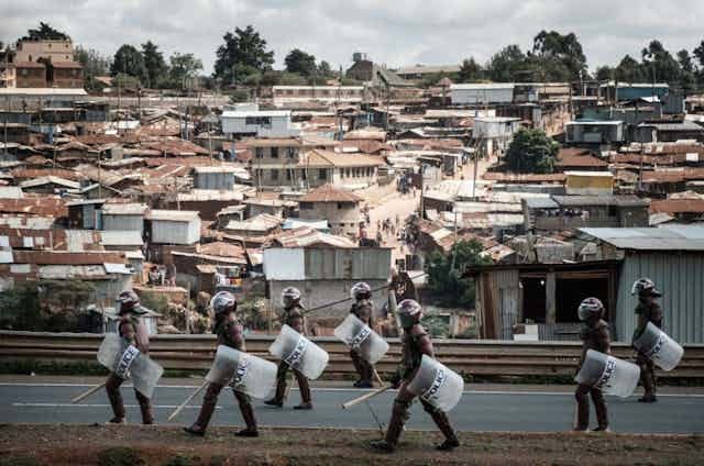 People in helmets, carrying truncheons and shields, walk on a road with a dense informal settlement in the background