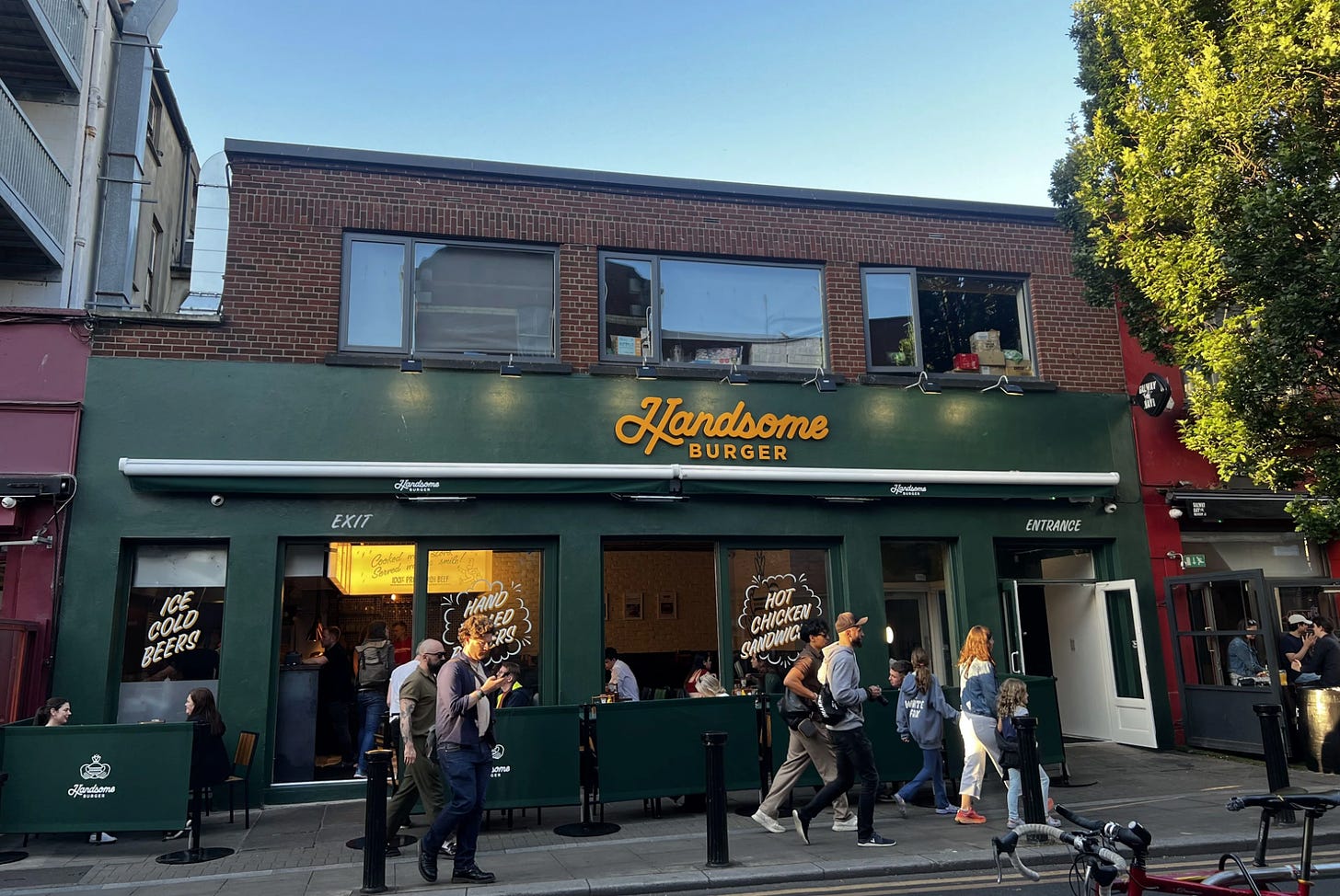 Dublin location: With a green face font and awning on the street, the distinctive Claddagh Ring logo alerts us that this is in fact Handsome Burger. Painted on the windows are slogans like Ice Cold Beers! Hot Chicken Sandwiches! It's a busy summer's day, with people eating inside and outside.