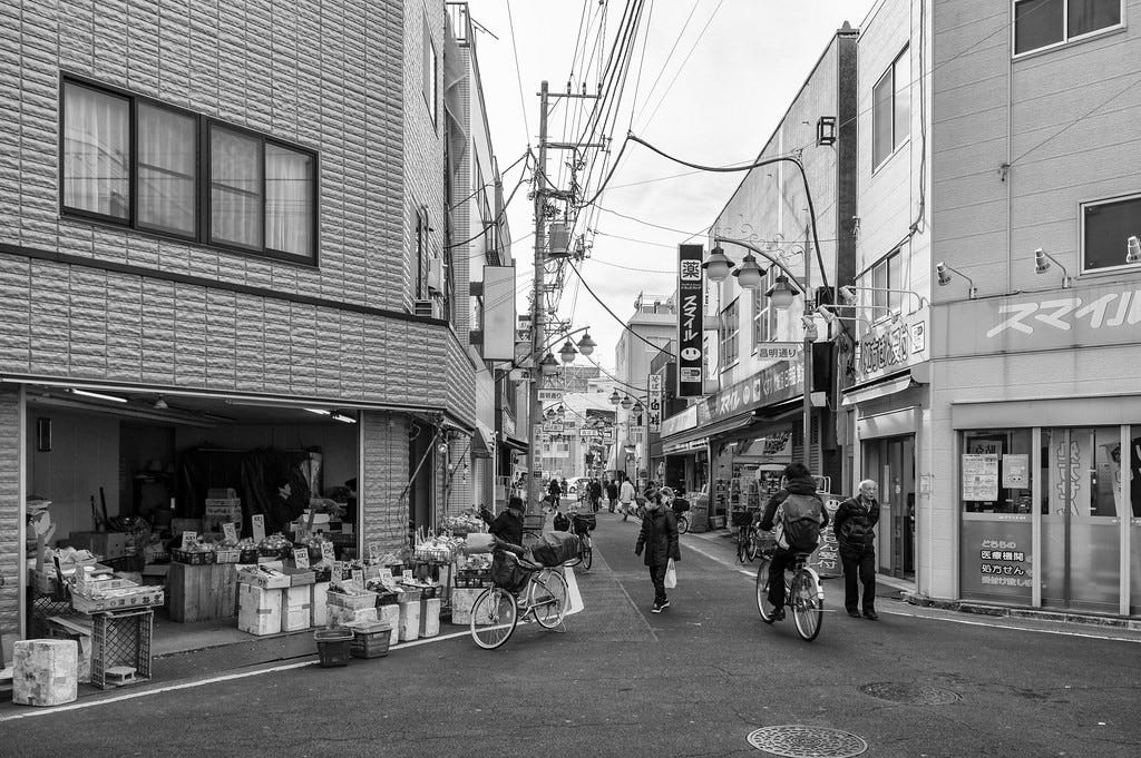 Monochrome image of a street in Higashi-Kanamachi, Tokyo, in winter. To the left foreground is a produce store with an open roll-up door. In the center foreground are a bicyclist, a pedestrian with a shopping bag, and a stooped old man. As the street recedes, we see power lines overhead, store signs and storefronts, and more pedestrians toward the end of the street.