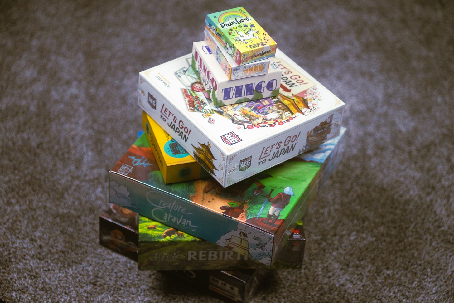 A stack of board games on the floor, including Rainbow, Prey, Tango, Let's Go! To Japan, Typewriter, Creature Caravan, Rebirth and Arcs.