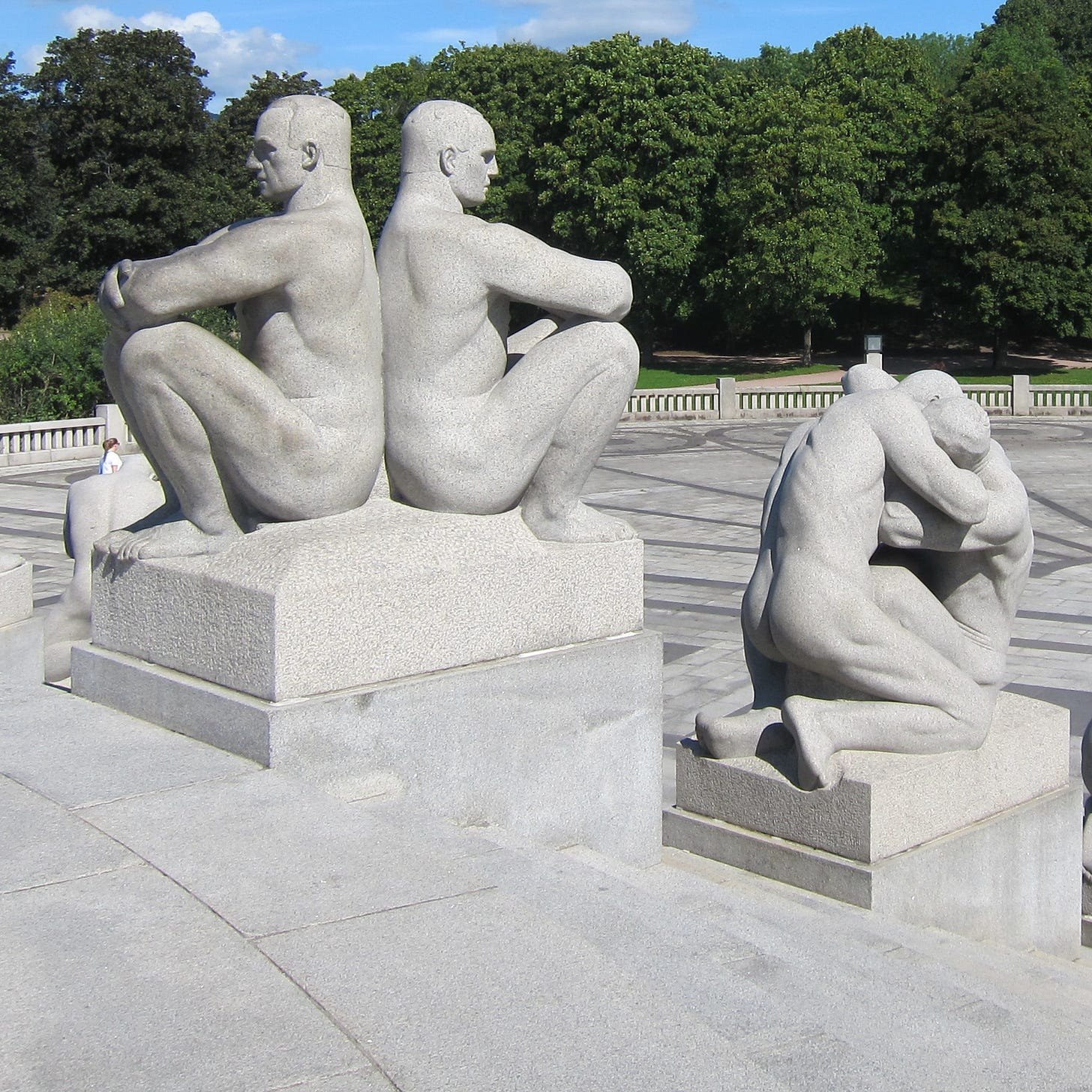 one statue with 2 people sitting back to back with arms crossed angrily and another statue of 2 people embracing with blue sky and green trees in background