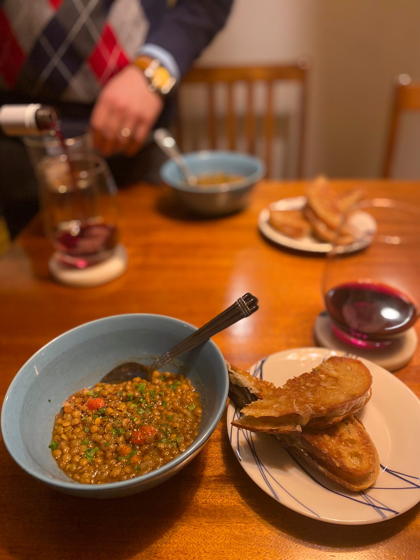 A blue bowl of chunky lentil soup next to a small plate with a grilled cheese sandwich. In the background, Jeff pours red wine into a stemless glass.
