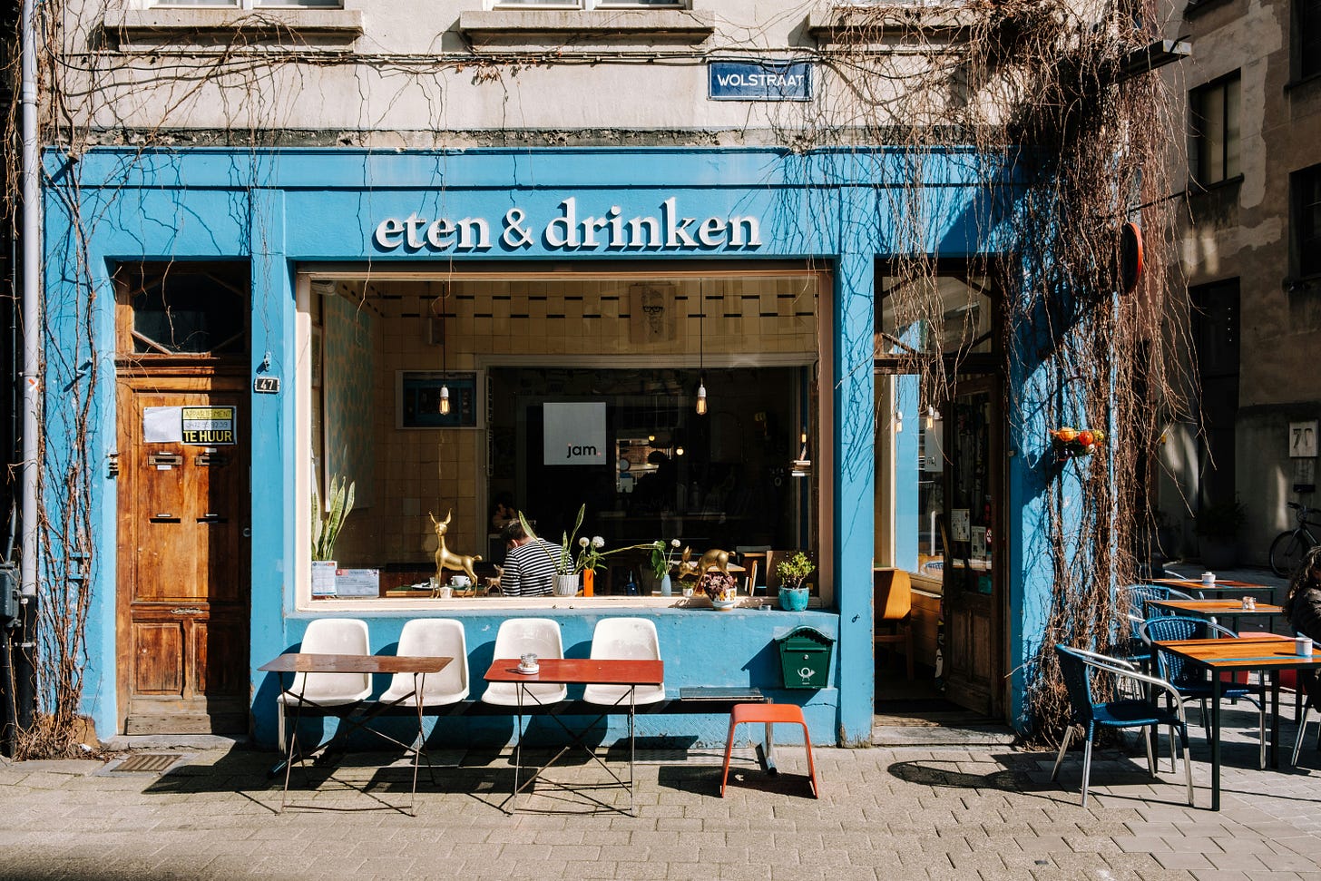 a cafe storefront in the sunshine. the shop has light blue walls and trim with a wooden door. four white plastic chairs sit out front facing away from the storefront, with trays in front of them for food. three wooden tables with green metal chairs sit close by. the sign says 'eten & drinken'