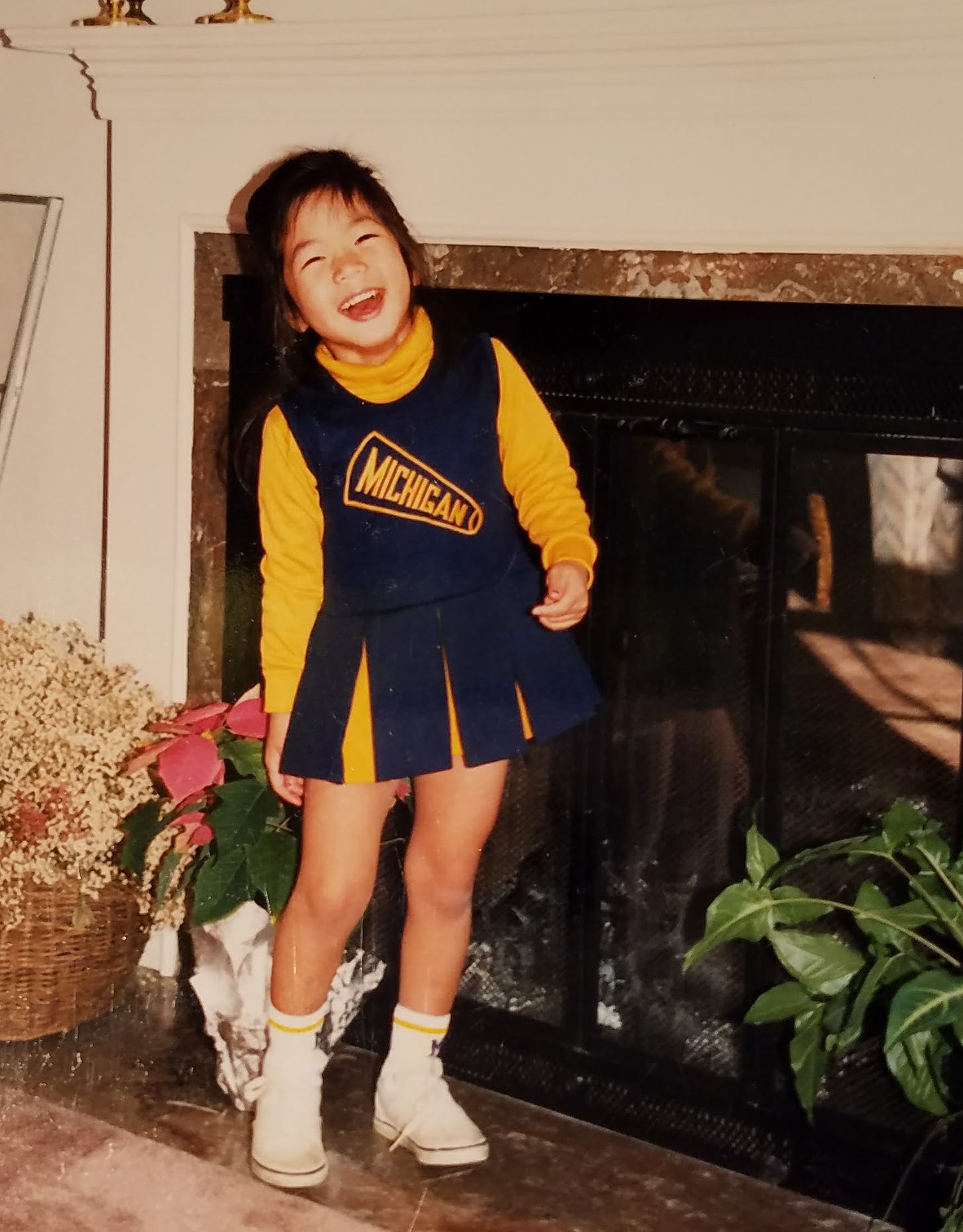 A Korean Adoptee stands next to a fireplace in a grainy photo. They wear a michigan cheerleader outfit and look joyous.