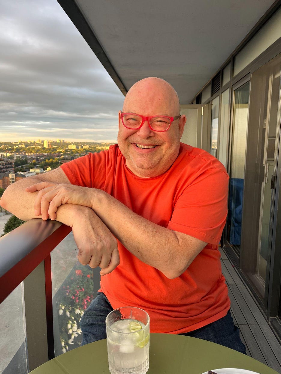 Kirby wearing pink glasses and a orange shirt sits on a balcony with a gin and tonic.