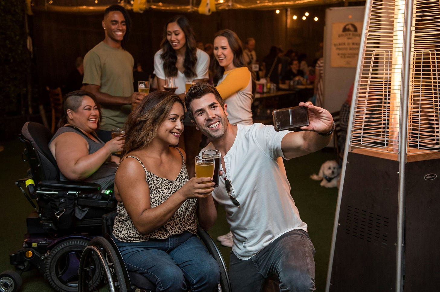 A white man in a wheelchair kneels down to take a selfie with a brown woman in a wheelchair as they clink drinks. Behind them, another woman in a wheelchair holding a drink laughs, and behind her, three non-visibly disabled people laugh too. 