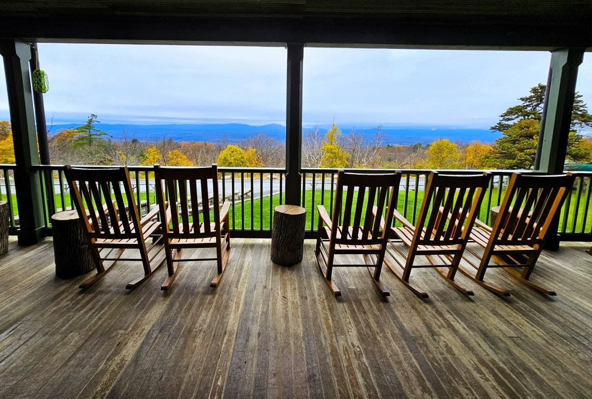 Five wooden rocking chairs are lined up on a wooden porch with a vast view of mountains and trees.
