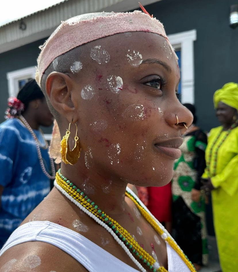 Juju, initiated as a priestess devoted to the orisha Oshun. she has red and white dots on her face, gold earring and nose jewelry, and yellow beads around her neck. picture taken in profile.
