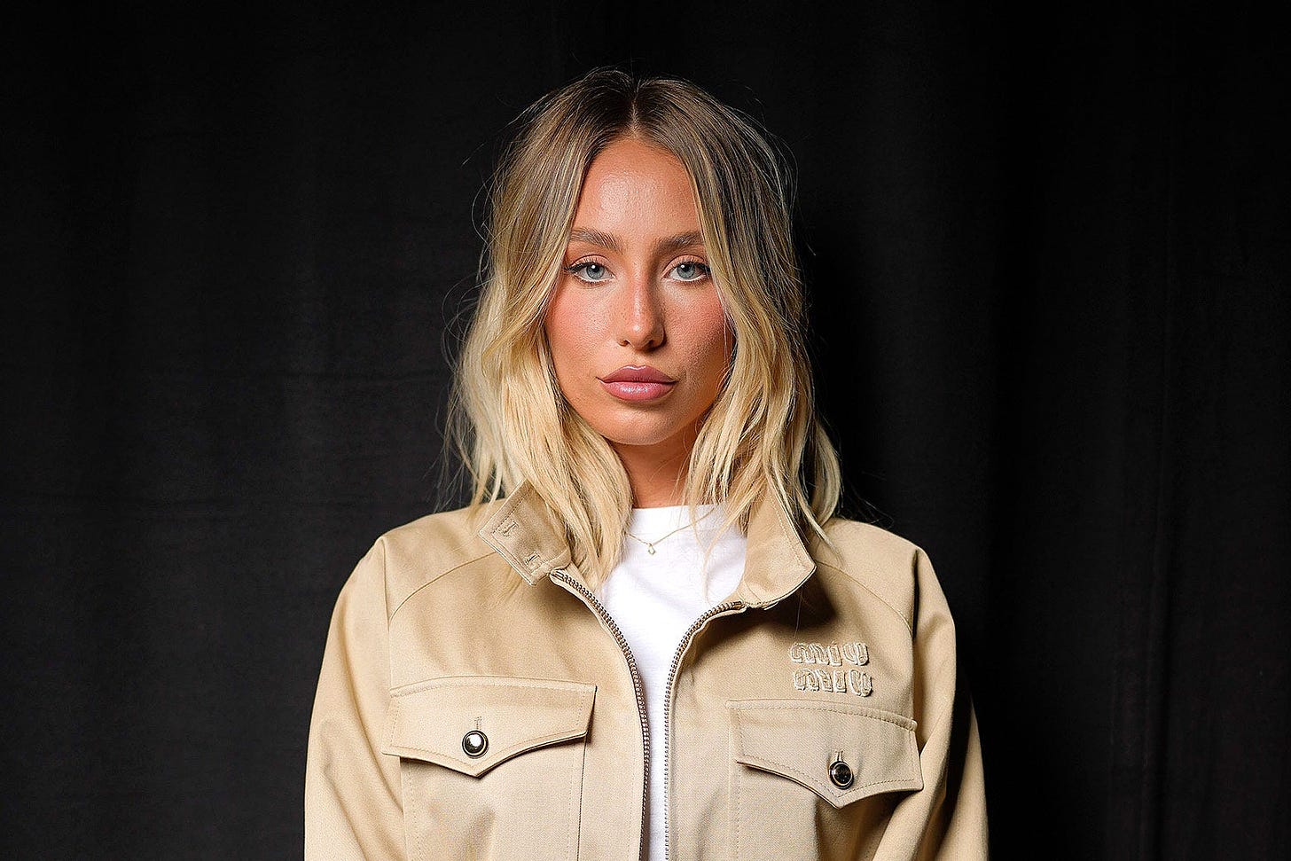 A blond white woman in a white shirt and tan jacket slightly smirks and looks ahead, in front of a black backdrop.