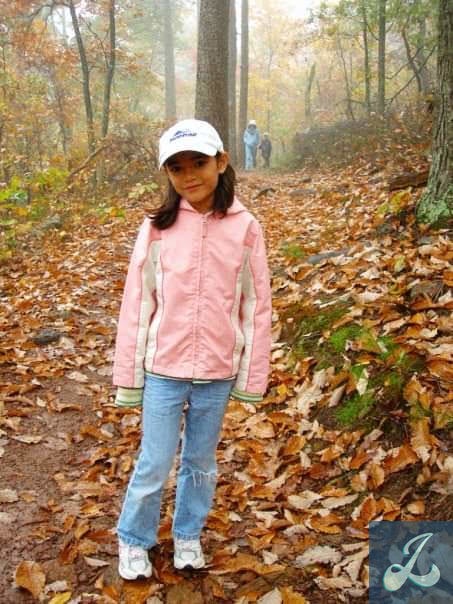 A girl is standing on a hiking trail covered in autumn leaves. She is wearing a pink windbreaker jacket; blue jeans with a rip in the left knee, white sneakers, and a white ballcap. She has tan skin, brown eyes, and dark brown hair.